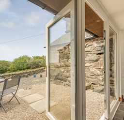 A glass door opening to a gravel patio with a metal table and chairs and a stone wall at Y Becws in Llanbedr near Harlech