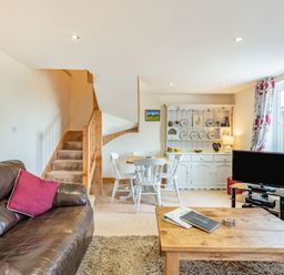 A living room with a brown leather sofa a wooden coffee table a dining table with white chairs a white cabinet a TV and stairs at Harvest Moon in Grundisburgh