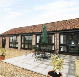 A patio area with a round table, chairs, and a closed umbrella outside a brick building with tiled roof at Jacko's Barn in Homersfield