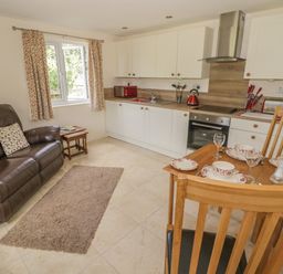 A kitchen and living area with a brown leather sofa wooden dining table set with dishes and glassware and white cabinets at Lawn Lodge in Ashleworth