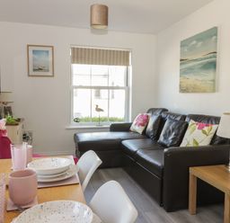 A living room with a brown leather sofa floral cushions a wooden side table with a lamp and a dining table set with plates cups and pink flowers at White Rose Apartment in Filey