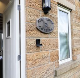 An open white door on a stone wall with a sign reading Kingfisher 3 Cottage and a window with blinds at Kingfisher in Whitby