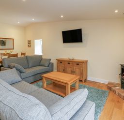 A living room with two gray sofas a wooden coffee table a wood-burning stove a wall-mounted TV a wooden sideboard and a dining table with chairs at Y Felin - Celyn in Borth