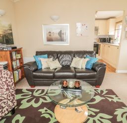 A living room with a dark brown leather sofa and a patterned armchair near a glass coffee table at Little Gem Lodge Charlesfield near St Boswells
