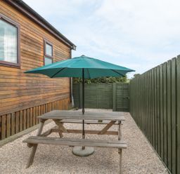 An outdoor area with a wooden picnic table and green umbrella next to a wooden building and green fence at Little Gem Lodge in Charlesfield near St Boswells