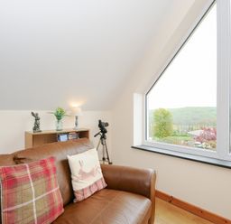 A leather sofa with two cushions next to a window with a countryside view and a small bookshelf with a lamp and decorative items at Treview in Launceston