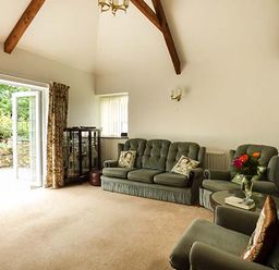 A living room with green upholstered sofas and armchairs a glass cabinet and open patio doors at Shepherds Rest in Jacobstow near Bude