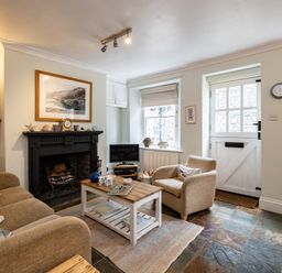 A living room with a beige sofa and armchair a wooden coffee table a black fireplace and a white door at Cobble Cottage in Boscastle near Tintagel
