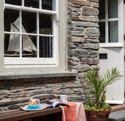 A stone exterior wall with a window displaying a model sailboat a white door a bench with a hat a magazine a mug and a tablecloth and a potted plant at Cobble Cottage Boscastle near Tintagel