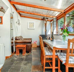A dining area with wooden framed chairs and a table with flowers and wine glasses a leather armchair and a stove in the corner at Hwylfa'r Groes in Talybont near Barmouth