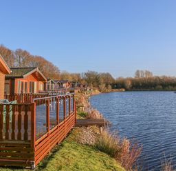 A row of wooden cabins with decks overlooking a body of water and trees at Shore Beats Work - Capenwray 10 in South Lakeland Leisure Village