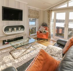 A living room with a leather sofa with cushions a glass coffee table an orange armchair a wall-mounted TV and a fireplace with candles at Shore Beats Work - Capenwray 10 in South Lakeland Leisure Village