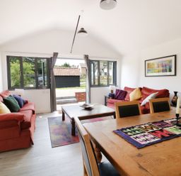 A living room with red sofas and colorful cushions near large windows and wooden dining table with candles at Whitehands Farm Barn in Old Buckenham