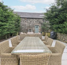 An outdoor patio with a glass top dining table and wicker chairs surrounded by tall trees at Yew Cottage in Backbarrow