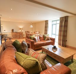 A living room with brown leather sofas green and plaid cushions wooden coffee table dining table and chairs with plaid curtains at Yew Cottage Backbarrow