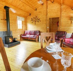 A wooden living room with red leather sofas a wood burning stove and a dining table with glassware at Acorn Lodge in Saxmundham