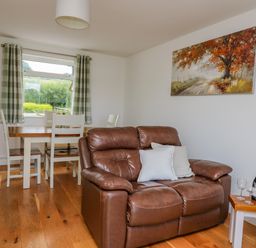 A brown leather sofa with pillows a small table with wine bottle and glasses a dining table with chairs and a window with curtains in a room at Foxes Meadow in Howey near Llandrindod Wells
