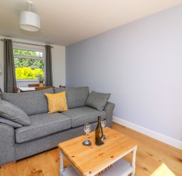 A living room with a grey sofa a yellow cushion a wooden coffee table with a bottle and two glasses and a dining table with chairs near a window at Owl Cottage in Llandrindod Wells