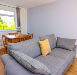 A gray sofa with a yellow pillow in a room with a wooden dining table and chairs near a window with gray curtains at Owl Cottage in Llandrindod Wells