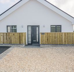 A front view of a single story house with a black door and two windows with wooden fences on both sides and gravel driveway at The Beach Halt in Perranporth