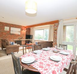 A dining table with red floral tablecloth and set plates and glasses in a living room with brown sofas and brick walls at Swan Dyke Cottage in Horning near Wroxham