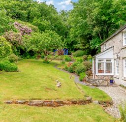 A stone cottage with a white porch next to a garden with trees shrubs and a lawn at Westside Cottage in Cardinham near Bodmin