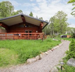 A wooden cabin with a gravel path leading to it surrounded by grass and trees at Heron Lodge in North Kessock
