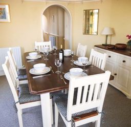 A dining room with a wooden table set for six with white chairs and tableware at Pendeen in Wadebridge