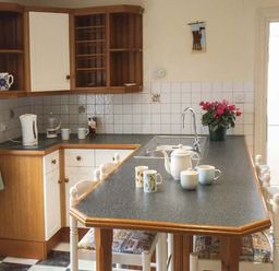 A kitchen with a black countertop island with cups and a teapot on it wooden cabinets a sink and a window with curtains at Pendeen in Wadebridge