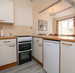 A kitchen with white cabinets a small oven a fridge and a dining area with chairs and a window at The Cottage in Chilsworthy near Holsworthy