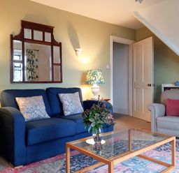 A living room with a blue sofa two patterned cushions a grey armchair with a red cushion a wooden glass coffee table with a vase of flowers and two side tables with lamps at Haslington House in Malvern