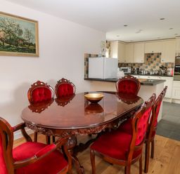 A dining room with a wooden table and red upholstered chairs next to a kitchen with cream cabinets and tiled floor at Honeysuckle Cottage Ashwater near Holsworthy