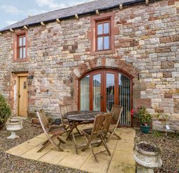 An outdoor patio with a round wooden table and four wooden chairs in front of a stone building with arched glass doors and two windows at The Hayloft in Milton near Brampton