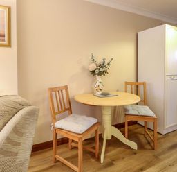 A small dining area with a round wooden table two wooden chairs with cushions a vase with flowers on the table a white cabinet and part of a beige armchair visible at Kingfisher in Wattisfield near Rickinghall and Diss