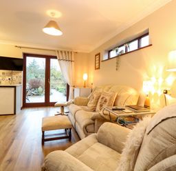 A living room with beige sofas a footstool a side table lamps a television kitchen appliances and glass doors leading outside at Kingfisher in Wattisfield near Rickinghall and Diss