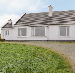 A single story white house with multiple windows a chimney a gravel driveway and grass in front at Seaspray in Tully County Galway