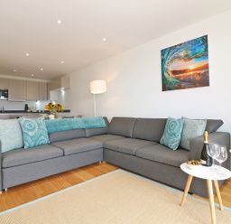 A living room with a gray sectional sofa and blue pillows next to a kitchen with cabinets at Saltwater One Lusty Glaze in Newquay