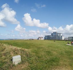 A large grassy field with a stone marker and modern buildings in the background at Saltwater One Lusty Glaze in Newquay
