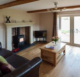 A living room with a wood burning stove a brown leather sofa a wooden coffee table with flowers and magazines and glass doors with curtains at Rose Cottage in Winforton near Hay-on-Wye