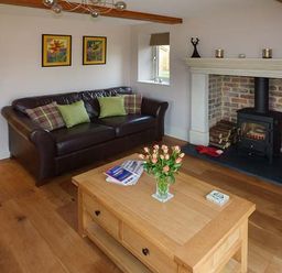 A living room with a brown leather sofa green and plaid cushions a wooden coffee table with flowers books and a fireplace with wood logs at Rose Cottage in Winforton near Hay-on-Wye