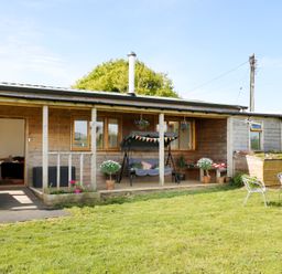 An outdoor view of a wooden cabin with a covered porch swing and potted plants in a grassy yard at View The Gallery in Okehampton