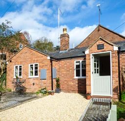 A brick cottage with a half open door a small patio with a table and chairs and a gravel driveway at Kitts Cottage in Little Malvern near Malvern