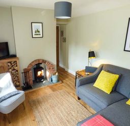 A living room with a fire in the stove a grey sofa with yellow cushions a wooden cabinet with firewood and a TV at Kitts Cottage in Little Malvern near Malvern