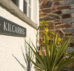 A black sign with Kilcarreg on a white wall next to a window and a green plant outside the building at Kilcarreg Rock near Wadebridge