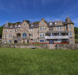 A large stone building with multiple windows and pitched roofs with turrets with a grassy lawn in front at Menai View near Menai Bridge