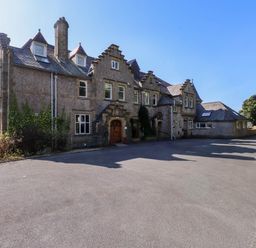 A stone building with multiple windows and chimneys with a paved driveway and parked cars near Menai Bridge