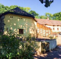 A thatched roof cottage with yellow walls and a glass extension surrounded by trees and plants at The Milking Parlour in Highampton