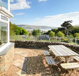 An outdoor patio with a wooden picnic table and benches overlooking a stone wall and greenery at Arnant in Barmouth