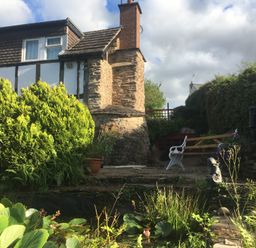 A garden with a stone chimney house bushes a bench a statue and a pond at Causeway Cottage in Pencombe