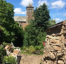 A garden bench on a stone path near a stone building with trees and a church tower in the background at Causeway Cottage in Pencombe